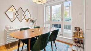 Een eetruimte met een houten tafel, groene beklede stoelen, een wit kastje met een geometrisch patroon en een blauw vloerkleed.