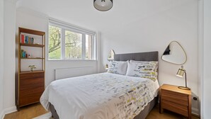 Een slaapkamer met een bed, nachtkastje, een boekenplank en een raam.
