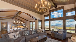 A spacious living room with a large sectional sofa, a wooden coffee table, and a view of mountains through large windows. - Mountainside Vista - Luxury Home with Stunning Views - Ski In / Ski Out! (Granby)
