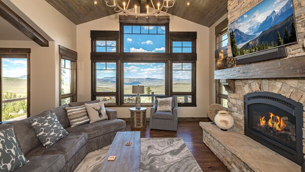 A spacious living room with a fireplace, large windows offering mountain views, a chandelier, and a stone fireplace surround. - Mountainside Vista - Luxury Home with Stunning Views - Ski In / Ski Out! (Granby)