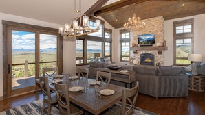 A spacious living area with a fireplace, a dining table set for six, and a large window offering a scenic view. - Mountainside Vista - Luxury Home with Stunning Views - Ski In / Ski Out! (Granby)