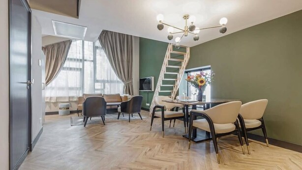 A modern living room with a herringbone floor, a dining area with a round table and chairs, a staircase, and a large window with curtains.