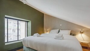 A modern hotel room with a large bed, a window with blinds, and a small bedside table with a lamp.