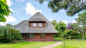 A house with a thatched roof and brick walls, surrounded by a well-maintained lawn and trees. - Watt. Familie. Reet. Objekt-id: Ejk/11/04 (Keitum)