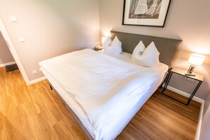 Een netjes opgemaakt bed met witte lakens en kussens, een nachtkastje met een lamp en een ingelijst foto aan de muur.