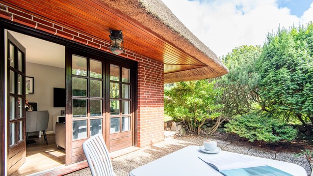 A patio with a table and chairs, a brick wall, and a glass door leading to an interior space. - Watt. Familie. Reet. Objekt-id: Ejk/11/04 (Keitum)