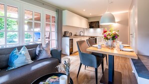 A modern kitchen with white cabinets, a dining table set for four, and a large window with a view of greenery. - Watt. Familie. Reet. Objekt-id: Ejk/11/04 (Keitum)