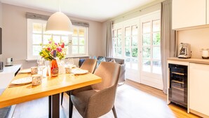 A modern kitchen with a dining area, featuring a wooden table, chairs, a pendant light, and a view of the outdoors through large windows. - Watt. Familie. Reet. Objekt-id: Ejk/11/04 (Keitum)