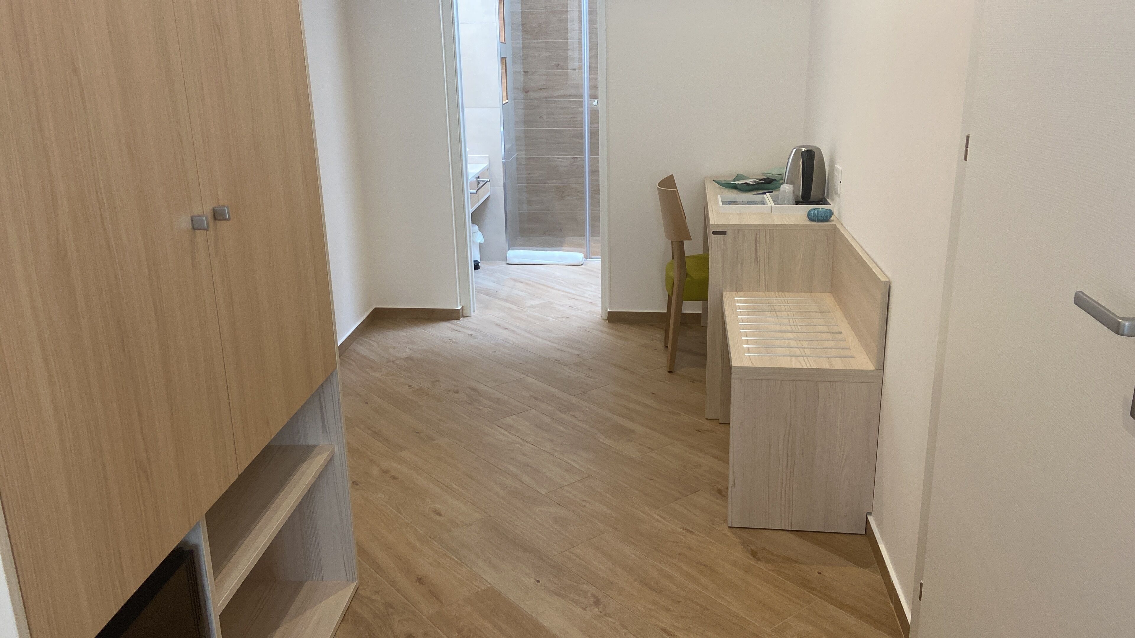 A hallway with wooden flooring, a small wooden bench, and a kitchenette area with a sink and a kettle.