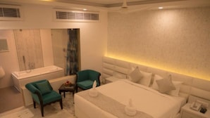 A modern hotel room with a large bed, a sitting area with a chair, a small table, a bathtub, and a vanity with a mirror. - Hotel Blue Waves (Port Blair)