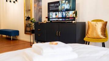 Habitación de hotel con cama, televisor reproduciendo una película, armario, banca y una silla dorada.