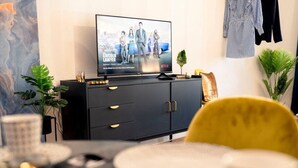 A modern living room with a flat-screen TV on a black cabinet, a dining table with plates and a yellow chair, and decorative plants. - Premium Studio | 1 Min to HBF (Mannheim)