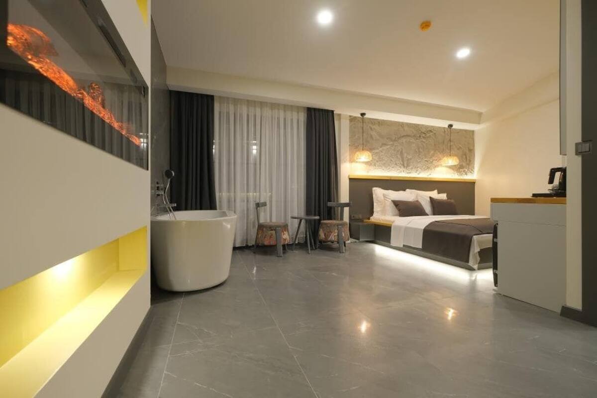 Una habitación de hotel moderna con una bañera grande, una cama con cabecera, una mesita con sillas y un ventanal con cortinas.