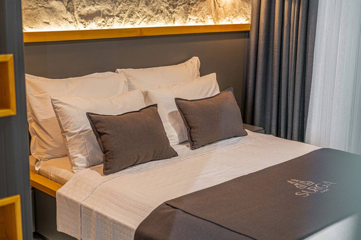 Una cama bien hecha con ropa de cama blanca y marrón, un cabecero y una pared con textura de fondo, y una mesita de noche oscura al lado de la cama.