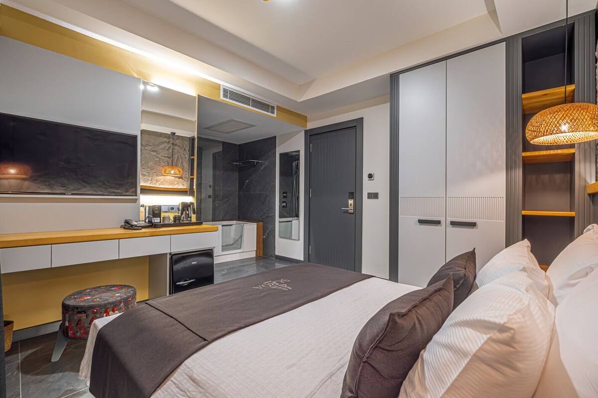 Una habitación de hotel moderna con una cama grande, un escritorio con computadora, un espejo, un armario y un baño con ducha e inodoro.