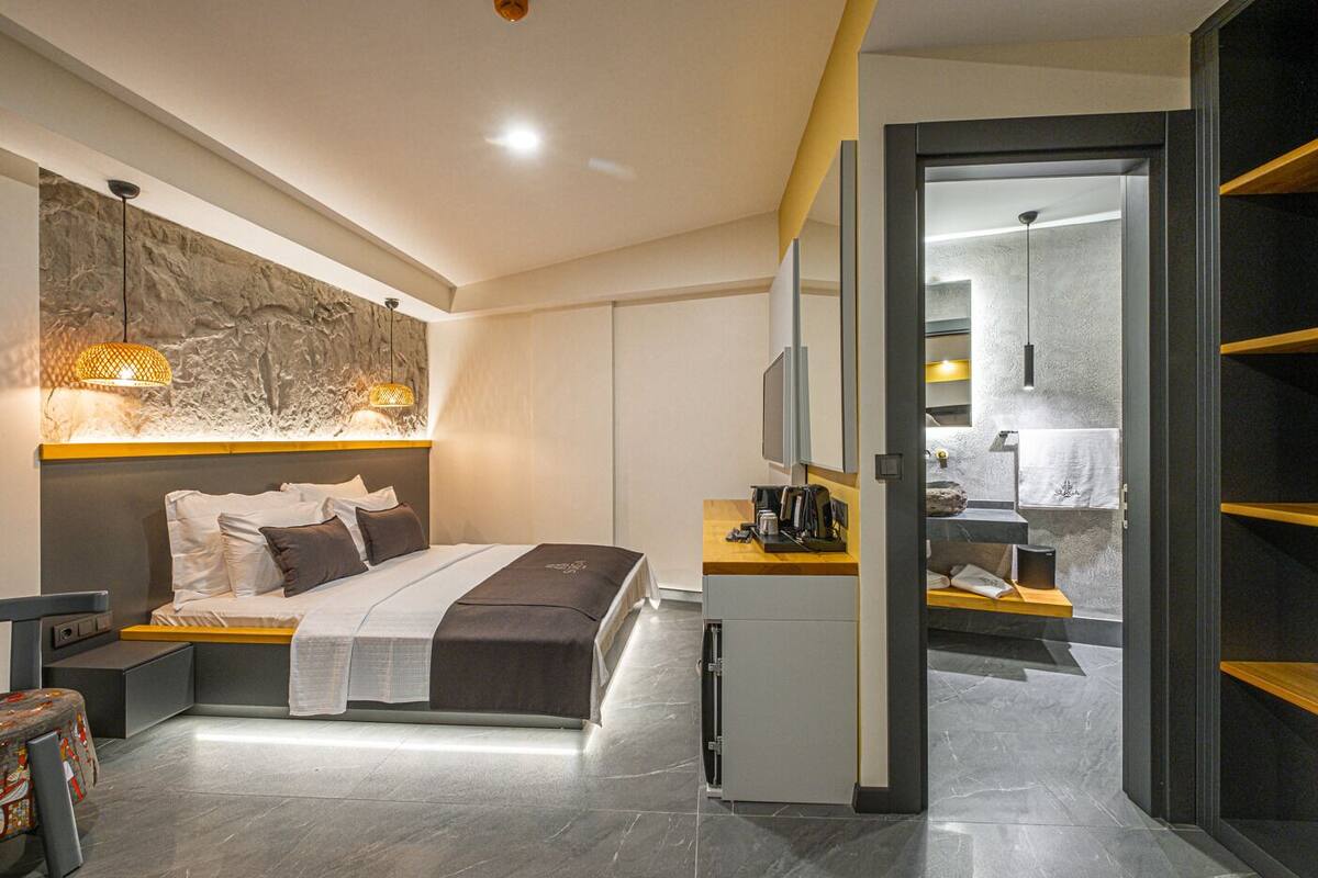 Una habitación de hotel moderna con cama, mesita de noche y un pequeño escritorio con silla. Hay un espejo grande sobre el escritorio y un televisor montado en la pared. La habitación tiene una pared de piedra y una puerta que conduce a un baño.