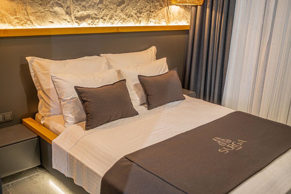 Una cama bien hecha con una manta y almohadas marrones, un cabecero con una lámpara y una cortina en la ventana.