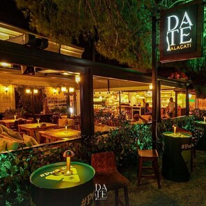 Un área de comedor al aire libre con mesas y sillas, un letrero con la inscripción 'DA TE ALACATI' y un cartel con un hashtag en un barril.
