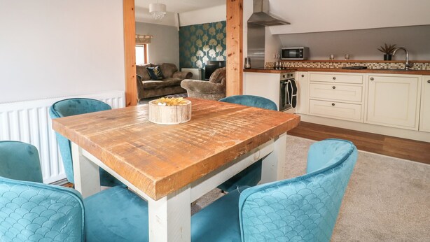 A dining area with a wooden table and blue chairs, a kitchen with white cabinets and a microwave, and a living room with a sofa and a patterned wall. - Old Print House (Ilkeston)