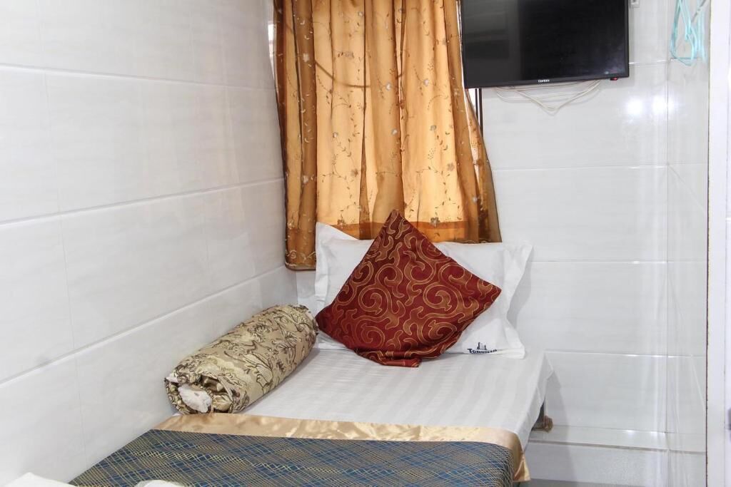 Una cama pequeña y bien hecha, con un cojín blanco y rojo estampado, una manta beige enrollada y un edredón de cuadros azules y beige.