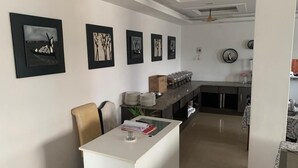 A modern kitchen with a central island, a dining area with a white table, and a hallway with framed artwork on the walls. - Airport Hotel (Port Blair)
