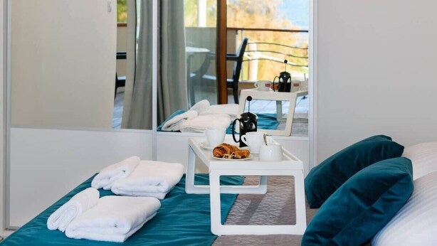 Une chambre d’hôtel avec un lit, une petite table sur laquelle se trouve un croissant, une tasse et une bouilloire. Il y a un miroir qui reflète la pièce, ainsi qu’un espace extérieur avec une chaise et une table.