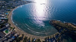 Vista aerea di una zona costiera con spiaggia, una piccola isola e un’area residenziale con edifici e piscina.