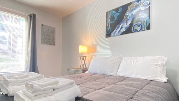 A neatly made bed with a grey duvet, white pillows, and folded white towels on the floor. There is a lamp on a bedside table and a painting on the wall. - 3rd Base Apt 1 - All Star Baseball Rentals (Oneonta)