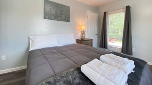 A bedroom with a large bed, a bedside table with a lamp, a window with curtains, and a view of greenery outside.