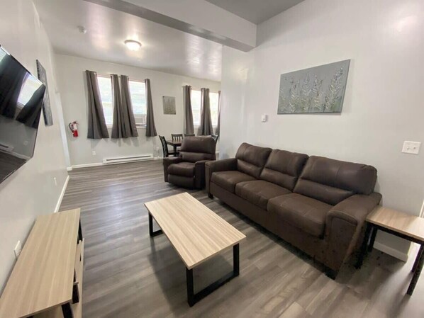 A modern living room with a brown leather sofa, two armchairs, a wooden coffee table, and a TV on the left wall. - Double Play Apt 1 - All Star Baseball Rentals (Oneonta)