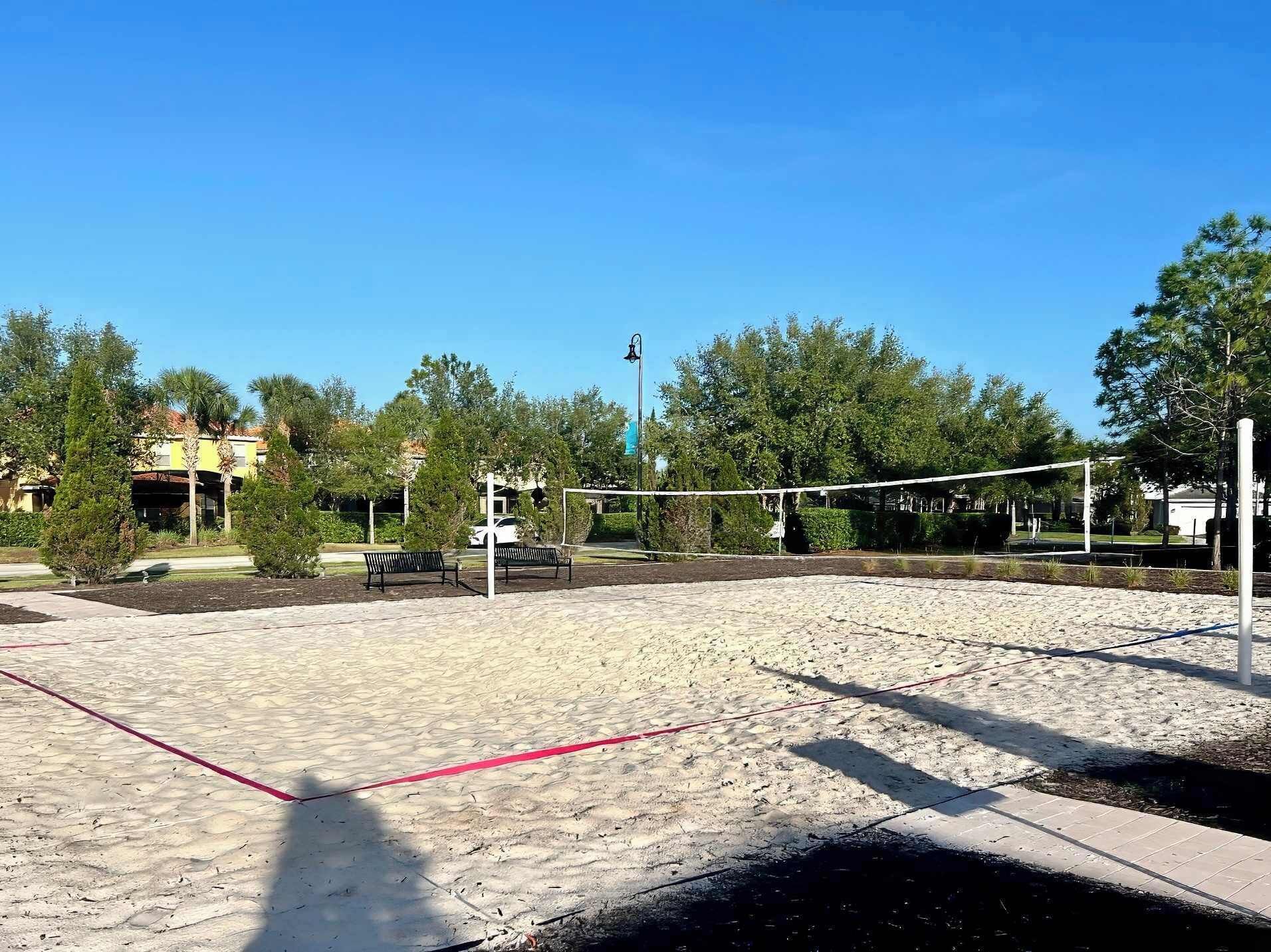 A volleyball court with a net, surrounded by trees and benches, with a building in the background.
