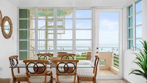 A dining area with a glass table and wicker chairs, large windows with a view of the sea, and a balcony with a railing. - Faro Penthouse | Beachfront with Stunning Views (Maunabo)