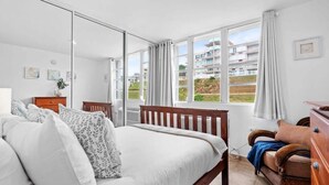 A bedroom with a large bed, a wooden dresser, a wicker chair, and a view of a residential area through large windows. - Faro Penthouse | Beachfront with Stunning Views (Maunabo)