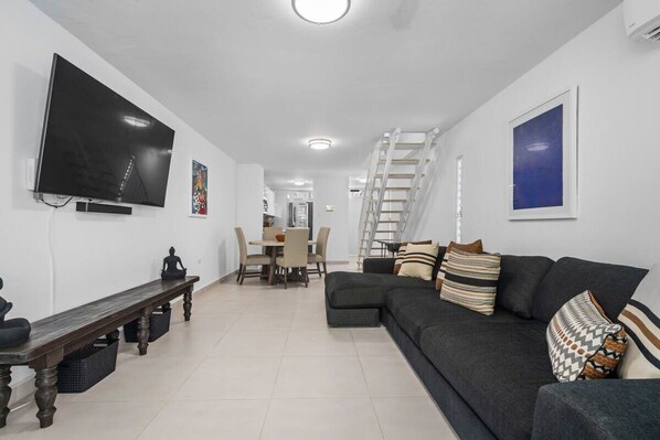 Una sala de estar moderna con un sofá gris oscuro, un televisor de pantalla plana montado en la pared, un banco de madera y un comedor con mesa y sillas.