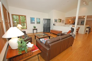 A modern living room with a brown leather sofa, a glass coffee table, and a wooden side table with a lamp and plant. A kitchen is visible in the background. - Dune Path - with Waterfront! (Empire)
