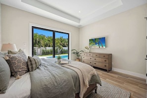 A bedroom with a large bed, a dresser, a painting, and a view of a pool and greenery.