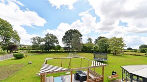 Une terrasse en bois avec une table et des chaises, un espace barbecue et une balançoire dans un jardin arrière verdoyant.