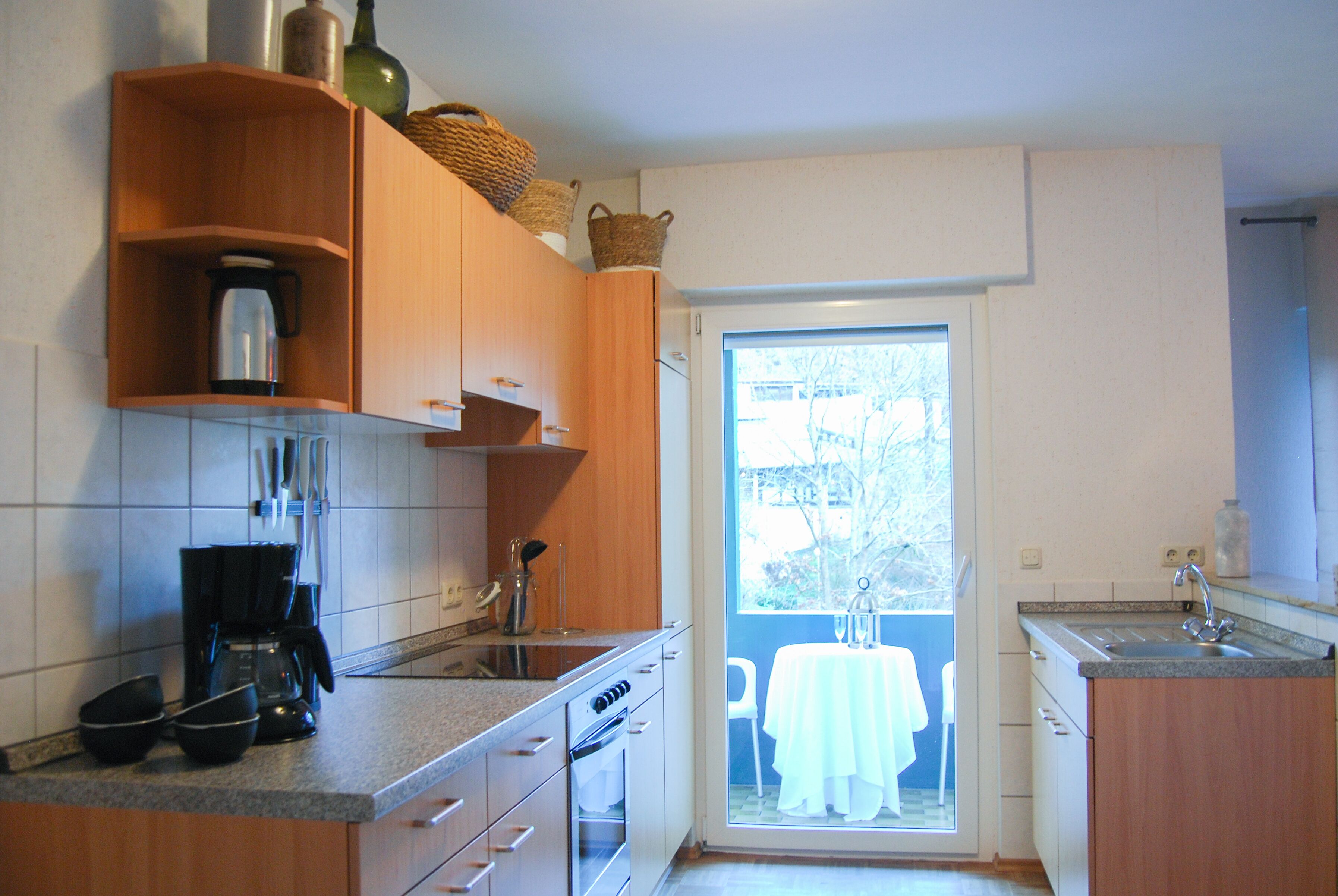 A kitchen with wooden cabinets, a coffee maker, and a view of a balcony with a white table and chairs.