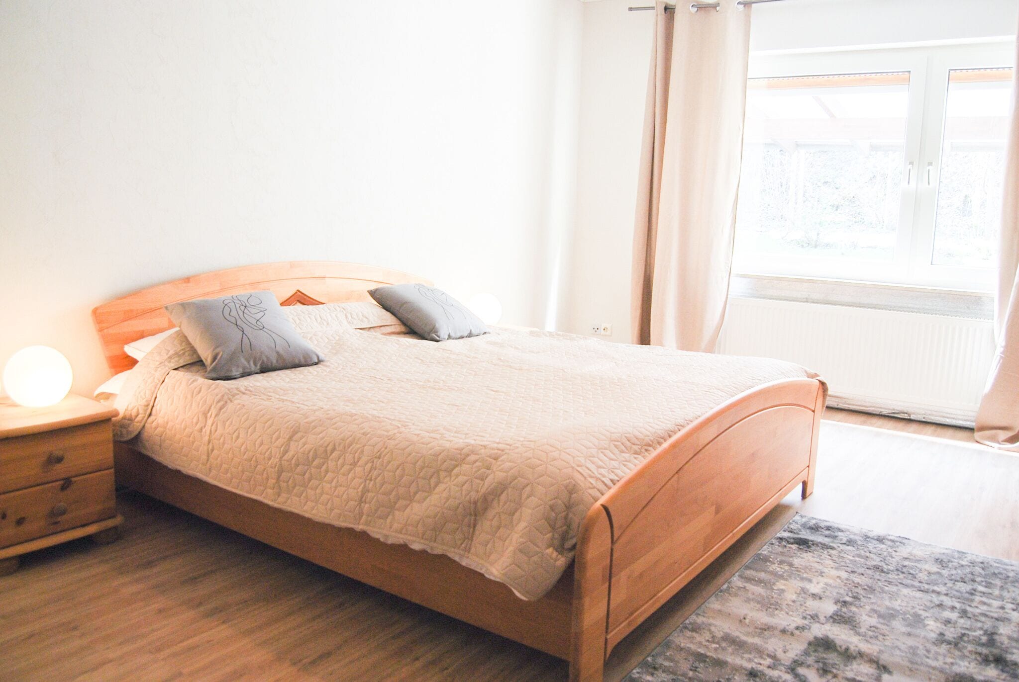 A neatly made bed with a wooden headboard, a bedside table with a lamp, and a view of the outdoors through a window with curtains.