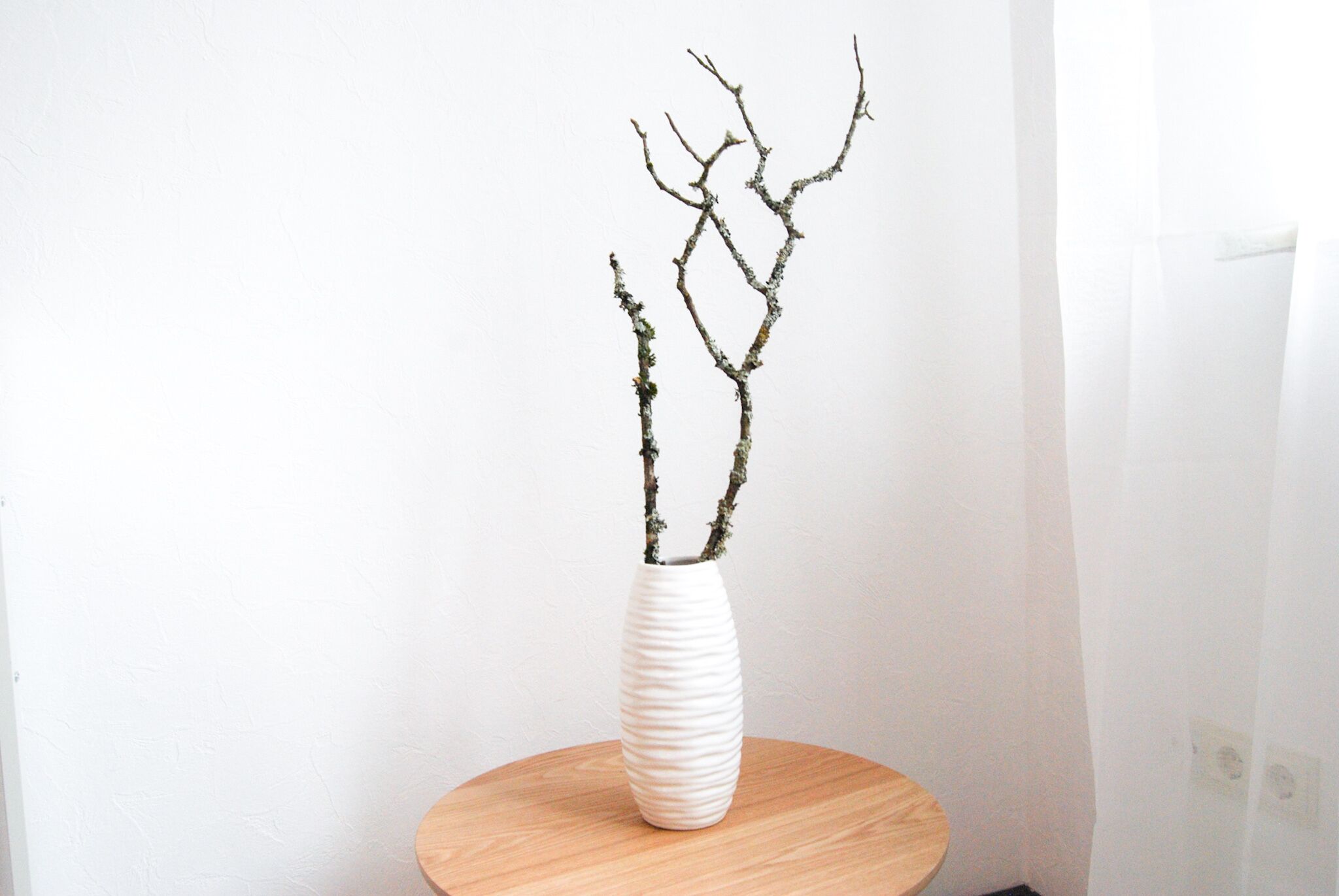 Un vase blanc au motif texturé, posé sur une table ronde en bois, devant un mur uni.