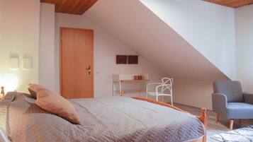 Een slaapkamer met een schuine plafond, een bed met een gewatteerde deken, een houten deur, een witte stoel en een grijze fauteuil.