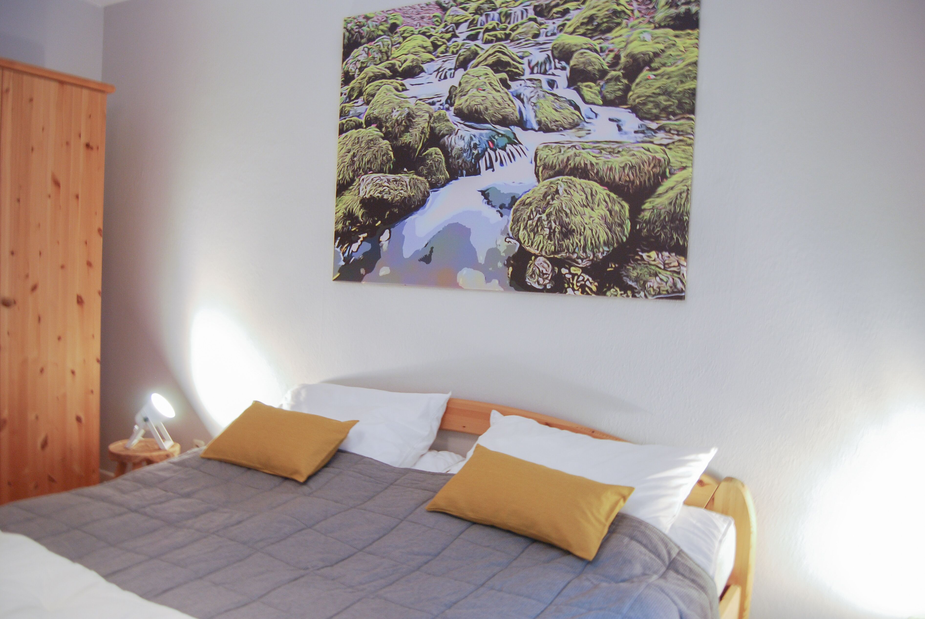 Ein Schlafzimmer mit einem Bett, zwei Kissen, einem Nachttisch, einer Lampe und einem Wandbild einer Landschaft.