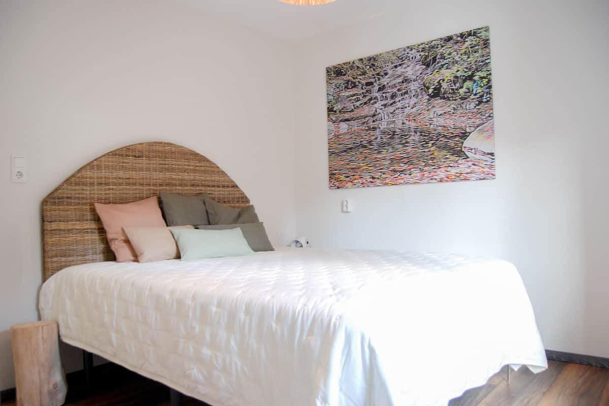 Une chambre à coucher avec un lit, des oreillers, une tête de lit, un tableau accroché au mur et un plancher en bois.