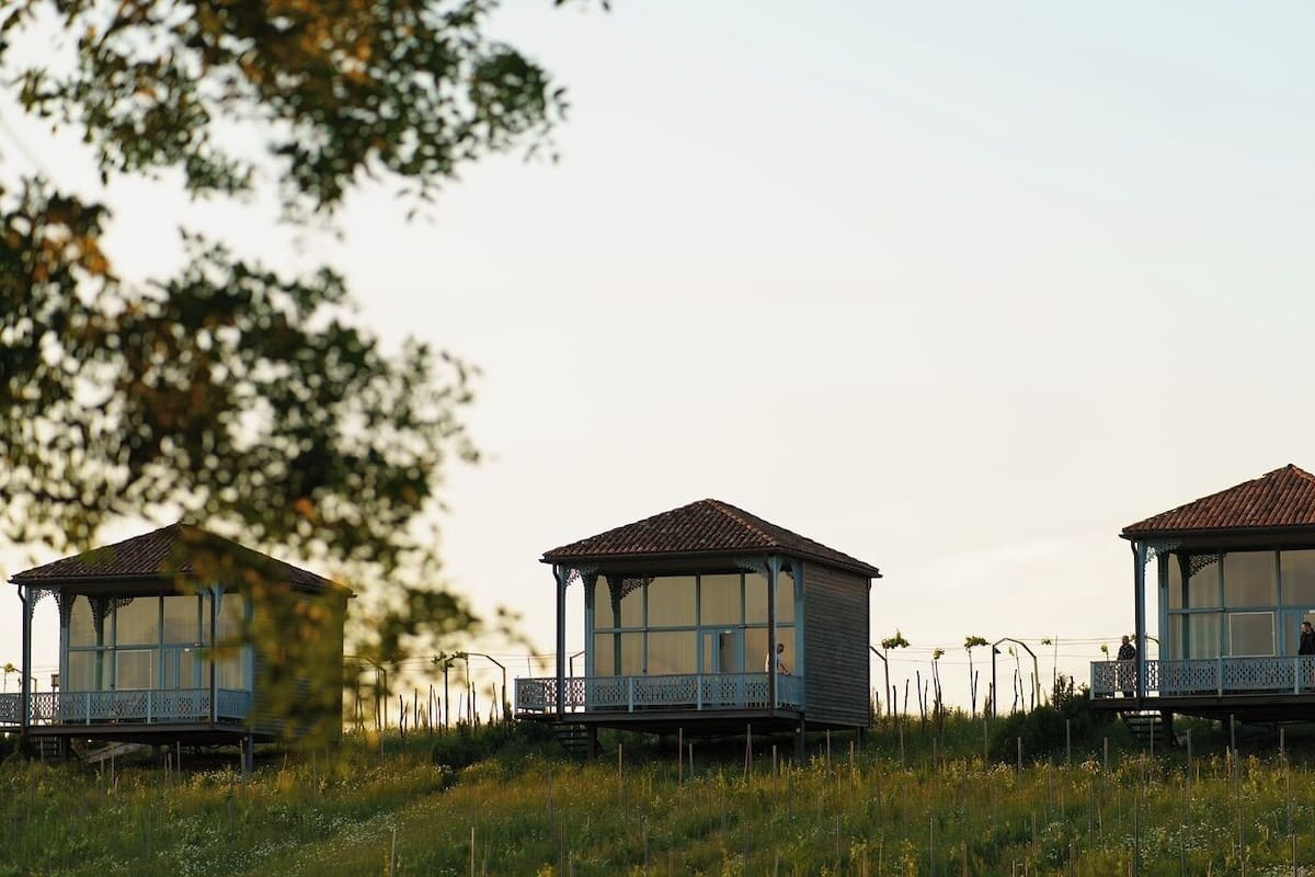 Three wooden gazebos with glass windows and balconies, situated in a grassy field with trees in the background.