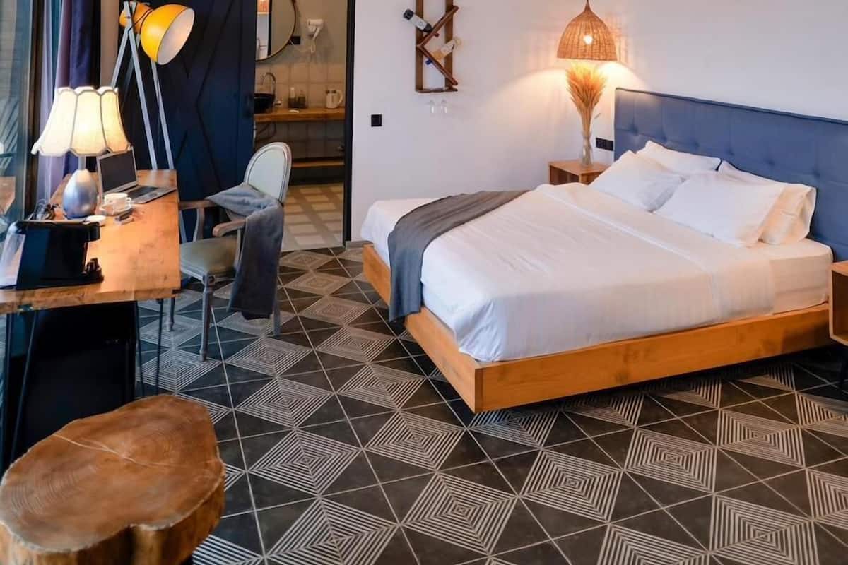 A modern hotel room with a geometric patterned floor, a wooden bed, a desk with a lamp, and two hanging pendant lights.