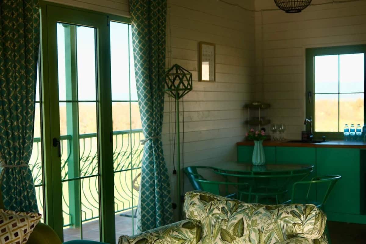 A room with a wooden table, a patterned sofa, a green dining area, and a window with a view.