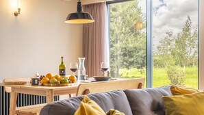 Una sala de estar moderna con un sofá, una mesa de comedor con frutas y copas de vino, y un ventanal con vistas a zonas verdes.