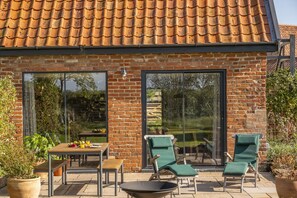 Een bakstenen huis met pannendak, grote glazen deuren, een terras met een tafel, stoelen en een vuurkorf.