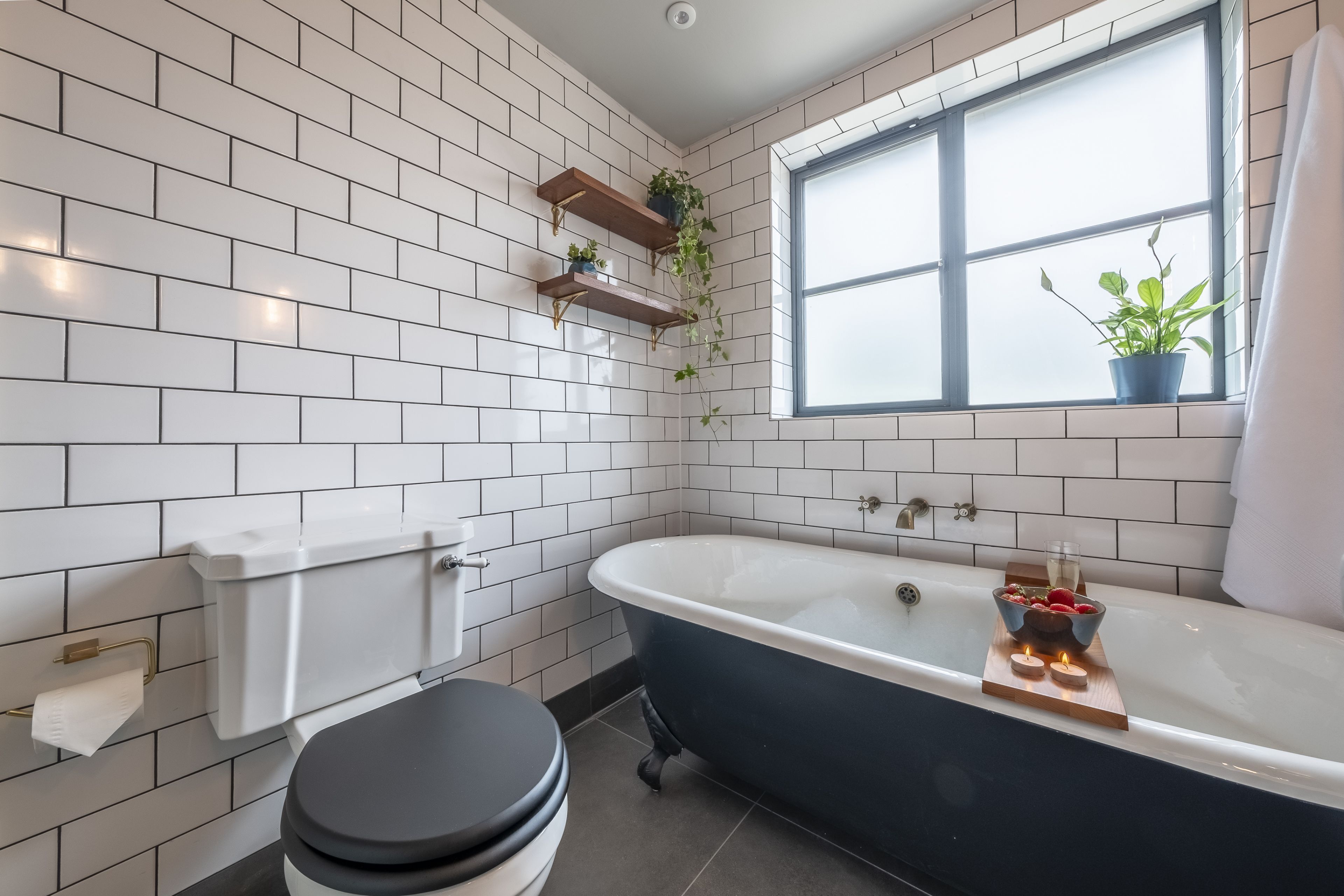 Un baño con paredes de azulejos blancos, una bañera negra, un inodoro y una ventana con plantas.