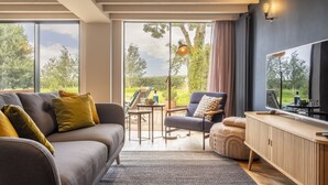 Una sala de estar moderna con sofá, sillón, reposapiés y mueble de televisión de madera. Amplias ventanas ofrecen vistas al exterior.